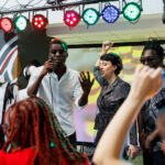 Band performing on stage with spotlights while crowd cheering and clubbing on nightclub dancefloor. Musicians singing and playing at electronic music festival live concert in club